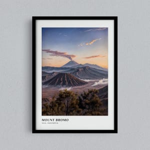 May include: A framed print depicting Mount Bromo in Java, Indonesia. The image shows a volcanic landscape with several mountains, including an active volcano with smoke. The sky transitions from blue to orange, with clouds. The print is in a black frame.