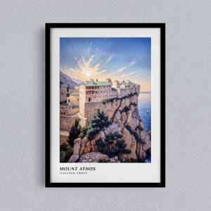 May include: A framed art print depicting a stone monastery perched on a cliffside overlooking the sea at Mount Athos, Chalkidiki, Greece. The scene features a sunrise sky, rocky terrain, and green trees. The text reads MOUNT ATHOS CHALKIDIKI, GREECE.