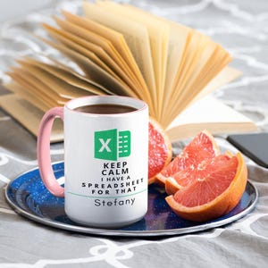 Puede incluir: Taza blanca con asa rosa, llena de café, y el texto "KEEP CALM I HAVE A SPREADSHEET FOR THAT" con el logo de Microsoft Excel. La taza está sobre un plato azul con rodajas de pomelo y un libro abierto.
