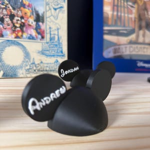 May include: Black 3D-printed mouse ear headbands with the names "Andrew" and "Jordan" in white script. The headbands are displayed on a light-coloured wooden surface, with Disney-themed artwork in the background.
