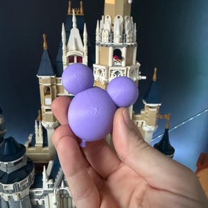 May include: A lilac Mickey Mouse-shaped balloon is held in front of a detailed model castle. The balloon has a textured surface and a small balloon knot. The castle features beige, white, and blue elements.