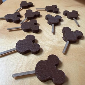 May include: Brown, Mickey Mouse-shaped treats on sticks, arranged on a light-coloured wooden surface. The treats have a textured appearance and are evenly spaced. The sticks are a light grey colour. The treats are likely for a party or celebration.
