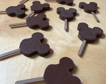 Puede incluir: Dulces marrones con forma de Mickey Mouse en palitos, dispuestos sobre una superficie de madera clara. Los dulces tienen un aspecto texturizado y están espaciados uniformemente. Los palitos son de color gris claro.