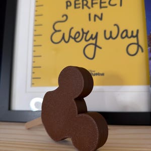 May include: A brown, 3D-printed Mickey Mouse-shaped popsicle on a wooden stick. The popsicle is resting on a wooden surface, with a framed yellow sign in the background that reads "Practically Perfect in Every Way."