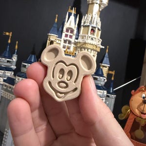 May include: A beige Mickey Mouse-shaped cookie cutter. The cutter is held in a hand, with a detailed castle model in the background. The cookie cutter features the iconic mouse's face with large ears and a smiling expression. The cutter is made of a light-coloured material.