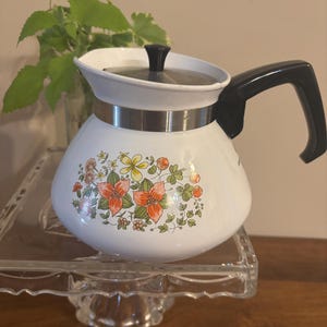 May include: A white enamel teapot featuring a floral pattern with orange, yellow, and green botanical illustrations. The teapot has a black handle, a silver-toned metal band around the upper section, and a matching white lid with a black knob.