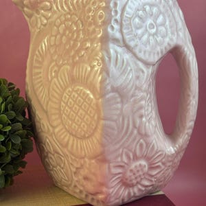 May include: A light pink ceramic pitcher with embossed floral designs. The pitcher has a curved handle and a spout for pouring. The floral patterns include sunflowers and other stylized flowers. The pitcher is sitting on a red book.