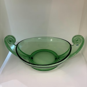 May include: A translucent light green glass bowl featuring a wide, circular base and two symmetrical, upward-curving handles on opposite sides. Each handle terminates in a decorative, tightly coiled spiral design at the top edge of the bowl.