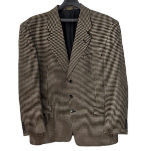 Könnte beinhalten: Ein Blazer mit braun-schwarzem Hahnentrittmuster. Das Kleidungsstück hat ein steigendes Revers, drei schwarze Knöpfe auf der Vorderseite, zwei Pattentaschen in der Taille und ein schwarzes Innenfutter. Die Ärmel haben kleine Knöpfe an den Manschetten.