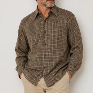 May include: A long-sleeve button-down shirt featuring a small-scale grid pattern in shades of brown, tan, and dark green. The shirt has a classic collar, a front button placket, and long sleeves with buttoned cuffs, worn over light tan trousers.