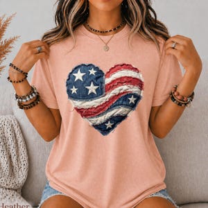 May include: Peach-colored t-shirt featuring a heart-shaped design in the style of the American flag. The heart is composed of denim blue fabric with white stars, and red and white striped fabric. A casual, patriotic apparel item.