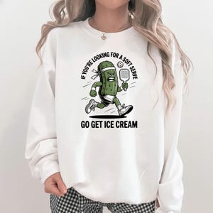 Könnte beinhalten: Weißes Sweatshirt mit einem Cartoon-Gurken-Charakter, der einen Schläger hält. Der Text auf dem Sweatshirt lautet: "IF YOU'RE LOOKING FOR A SOFT SERVE GO GET ICE CREAM". Die Gurke hat einen wütenden Gesichtsausdruck und rennt.