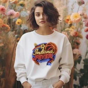 May include: A white crewneck sweatshirt featuring a graphic print on the chest. The design displays the text Indiana FEVER in a neon-style font, accompanied by a basketball, a hoop, and star shapes in shades of orange, red, blue, and yellow.