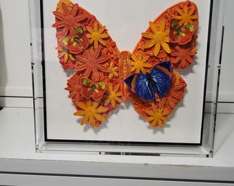 May include: A framed artwork featuring a vibrant butterfly design. The butterfly is composed of orange and yellow floral elements, with accents of red and blue. The artwork is encased in a clear acrylic box with a black border, creating a modern display.