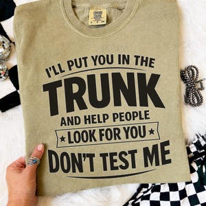 Może przedstawiać: Oliwkowo-zielona koszulka z napisem "I'll put you in the trunk and help people look for you, don't test me" w kolorze czarnym. Koszulka ma okrągły dekolt i krótkie rękawy. Koszulka jest wykonana z miękkiego materiału.