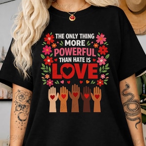 May include: Black t-shirt with the text "THE ONLY THING MORE POWERFUL THAN HATE IS LOVE" in red and green. The design includes floral elements and hands with hearts, promoting a message of love and unity.