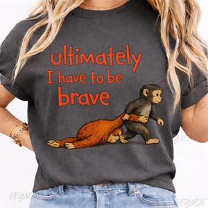 Op de afbeelding: Donkergrijs t-shirt met de tekst "Ultimately I have to be brave" in oranje. Het shirt heeft een cartoon illustratie van twee apen, waarvan één de ander trekt. Een casual kledingstuk.