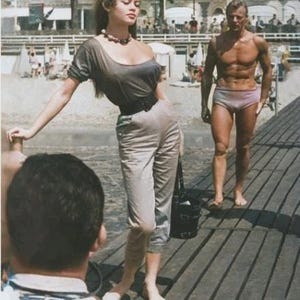 以下が含まれることがあります： ヴィンテージ写真。グレーのトップスとパンツ、黒いベルト、ビーズのネックレスを身につけた女性。水着姿の男性が木製の遊歩道に立っています。背景には建物とヤシの木があるビーチのシーンです。