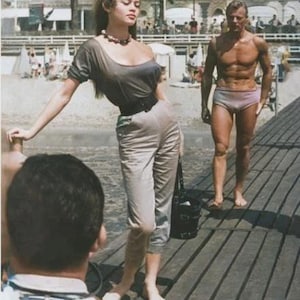 以下が含まれることがあります： ヴィンテージ写真。グレーのトップスとパンツ、黒いベルト、ビーズのネックレスを身につけた女性。水着姿の男性が近くの木の遊歩道に立っています。背景には建物とヤシの木があるビーチのシーンです。