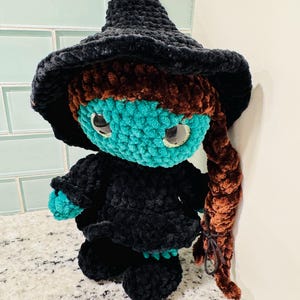 May include: A handmade crochet doll in the shape of a witch. The doll has a black hat and dress, teal skin, brown hair, and large, round eyes. The doll is made of soft, plush yarn.