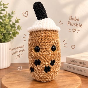 May include: A crocheted boba plushie featuring a tan cylindrical body with black circular accents, a white lid, and a black straw. The plushie has large black eyes and text that reads Boba Plushie and Handmade with love.