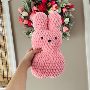 May include: A pink, plush bunny-shaped pillow with black button eyes and a black stitched mouth. The bunny is made of a soft, textured material. The background includes a floral wreath with pink and red flowers.