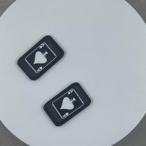 Può includere: Due toppe rettangolari nere con un disegno bianco di asso di picche. Le toppe sono su una superficie circolare bianca. Il disegno dell'asso di picche presenta una picca bianca, una tromba e i numeri "4" e "A".