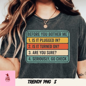 May include: Dark grey t-shirt featuring the text "BEFORE YOU BOTHER ME" and a troubleshooting checklist: "1. IS IT PLUGGED IN?", "2. IS IT TURNED ON?", "3. ARE YOU SURE?", and "4. SERIOUSLY, GO CHECK". The text is in a rectangular design with yellow, orange, and teal stripes.