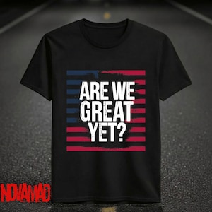 Op de afbeelding: Zwart T-shirt met de tekst "ARE WE GREAT YET?" in het wit, tegen een rood en blauw gestreepte achtergrond. Het shirt heeft een ronde hals en korte mouwen. De merknaam "NOVAMAD" staat in het rood onderaan.