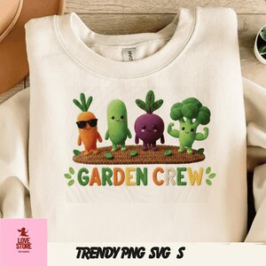 May include: Cream-colored sweatshirt featuring a colorful graphic of crocheted vegetables: a carrot, cucumber, beet, and broccoli, with the text "GARDEN CREW". Below the graphic is the text "TRENDY PNG SVG S".
