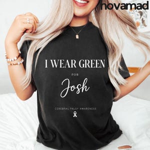 Op de afbeelding: Zwart T-shirt met de witte tekst "I WEAR GREEN for Josh". Het shirt bevat ook de tekst "CEREBRAL PALSY AWARENESS" en een witte lintafbeelding. De merknaam "novamad" staat rechtsboven.
