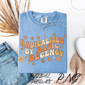 Op de afbeelding: Een lichtblauw T-shirt met de tekst "RADICALIZED BY BASIC DECENCY" in een retro lettertype in oranje en geel. Het shirt heeft bloemen- en vredestekenafbeeldingen. Een zilveren ketting met een hanger ligt op het shirt. De woorden "DIGITAL PRODUCT PNG" staan onderaan.
