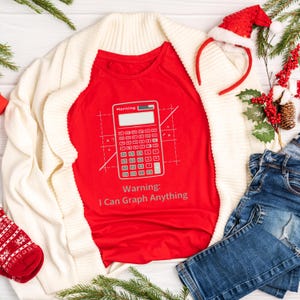 Puede incluir: Una camiseta roja con un gráfico de calculadora y el texto "Warning: I Can Graph Anything". La camiseta está rodeada por un cárdigan color crema, vaqueros azules, una diadema de gorro de Papá Noel y decoraciones navideñas.