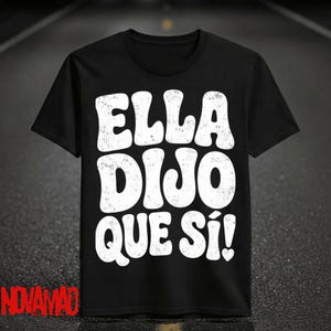 May include: Black t-shirt with the white text "ELLA DIJO QUE SÍ!" in a bold, distressed font. The shirt has a classic crew neck and short sleeves. The brand name "NOVAMAD" is printed in red at the bottom left.