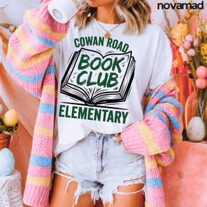 Può includere: T-shirt bianca con la scritta "COWAN ROAD BOOK CLUB ELEMENTARY" in verde e nero. La maglietta presenta una grafica di un libro aperto. Un maglione a righe rosa, giallo e blu è drappeggiato sulle spalle.