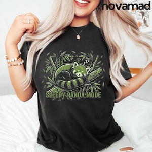 Może przedstawiać: Czarny t-shirt z zielono-czarną ilustracją śpiącej pandy rudej na gałęzi bambusa. Pod pandą znajduje się napis "SLEEPY PANDA MODE". Koszulka ma swobodny styl.