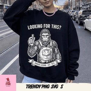 Könnte beinhalten: Schwarzes Sweatshirt mit dem Text "LOOKING FOR THIS?" über einer Grafik eines Gorillas in Latzhosen, der ein Werkzeug hält. Unter dem Gorilla steht ein Banner mit der Aufschrift "GARAGE LEGEND - EST. 1974".