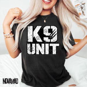 Könnte beinhalten: Ein schwarzes Kurzarm-T-Shirt mit einer großen, abgenutzten weißen Grafik auf der Brust, die K9 UNIT zeigt. Das Design enthält stilisierte Pfotenabdrücke in den Buchstaben und drei diagonale Kratzspuren über der Ziffer neun.