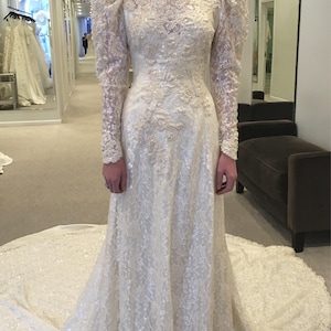 May include: Ivory wedding gown with a high neckline, long sleeves, and a full skirt with a long train. The dress features intricate lace and sequin detailing throughout. The sleeves have a puffed shoulder design.