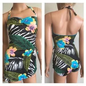 Pode incluir: Um fato de banho de uma pe&ccedil;a preto com padr&atilde;o de zebra, folhas verdes grandes e flores coloridas. O fato de banho tem um pesco&ccedil;o halter e uma saia envolvente. As flores s&atilde;o azuis, amarelas e rosas. Acabamento azul.