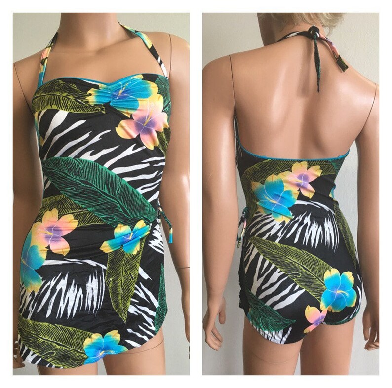 Pode incluir: Um fato de banho de uma pe&ccedil;a preto com padr&atilde;o de zebra, folhas verdes grandes e flores coloridas. O fato de banho tem um pesco&ccedil;o halter e uma saia envolvente. As flores s&atilde;o azuis, amarelas e rosas. Acabamento azul.