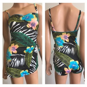 Pode incluir: Um fato de banho de uma pe&ccedil;a preto com padr&atilde;o de zebra, adornado com flores tropicais e folhas verdes. O fato de banho tem al&ccedil;as finas e uma saia envolvente. A borda &eacute; turquesa.