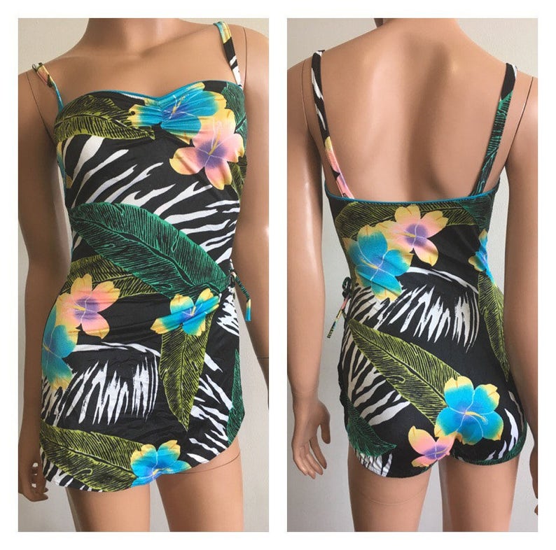 Pode incluir: Um fato de banho de uma pe&ccedil;a preto com padr&atilde;o de zebra, adornado com flores tropicais e folhas verdes. O fato de banho tem al&ccedil;as finas e uma saia envolvente. A borda &eacute; turquesa.