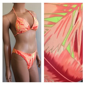 Pu&ograve; includere: Un bikini a due pezzi nei toni dell'arancione, del rosa e del giallo. Il top del bikini con collo all'americana e gli slip a vita alta presentano un motivo a foglie tropicali. Il costume da bagno &egrave; esposto su un manichino.