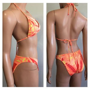 Pu&ograve; includere: Bikini a due pezzi con vivace stampa tropicale arancione, gialla e rossa. Il top all'americana e gli slip a vita alta presentano un motivo a foglie ripetuto. Il bikini &egrave; esposto su un manichino.