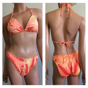 Pu&ograve; includere: Un bikini a due pezzi con un vivace motivo a foglie tropicali arancioni e gialle. Il top del bikini all'americana ha coppe a triangolo e lacci al collo e sulla schiena. La parte inferiore abbinata ha un design a taglio alto.