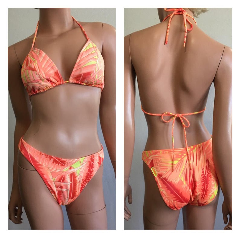 Pu&ograve; includere: Un bikini a due pezzi con un vivace motivo a foglie tropicali arancioni e gialle. Il top del bikini all'americana ha coppe a triangolo e lacci al collo e sulla schiena. La parte inferiore abbinata ha un design a taglio alto.