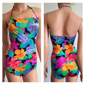 Peut inclure: Un maillot de bain une pi&egrave;ce avec un motif floral vibrant. Le maillot de bain pr&eacute;sente un col licou et un fond noir avec de grandes fleurs color&eacute;es dans les tons orange, rose, violet et bleu. Le design comprend des feuilles vertes.