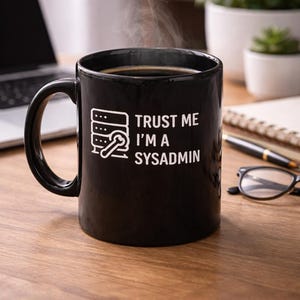 Könnte beinhalten: Eine schwarze Keramik-Tasse, gefüllt mit Kaffee, mit aufsteigendem Dampf. Auf der Tasse befindet sich eine weiße Grafik eines Servers und eines Schlüssels, sowie der Text "TRUST ME I'M A SYSADMIN". Die Tasse steht auf einer Holzoberfläche.