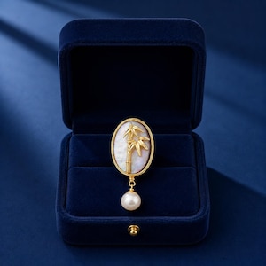 May include: A gold-toned brooch with an oval mother-of-pearl center featuring a bamboo design. A dangling pearl hangs below the brooch. The brooch is displayed in a dark blue velvet jewelry box.